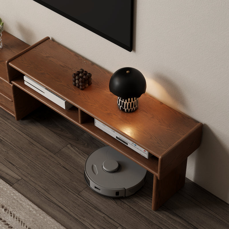 Wooden entertainment console with electronic devices and a black and white decorative item, with a robot vacuum cleaner in front.