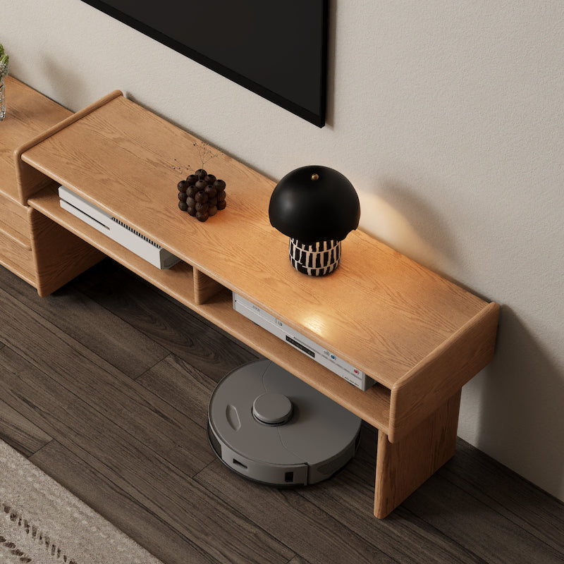 Wooden TV stand with electronic devices and a black and white decorative item, with a robot vacuum cleaner in front.