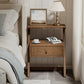 Wooden nightstand with a lamp, cup, and book next to a bed in a bedroom setting.