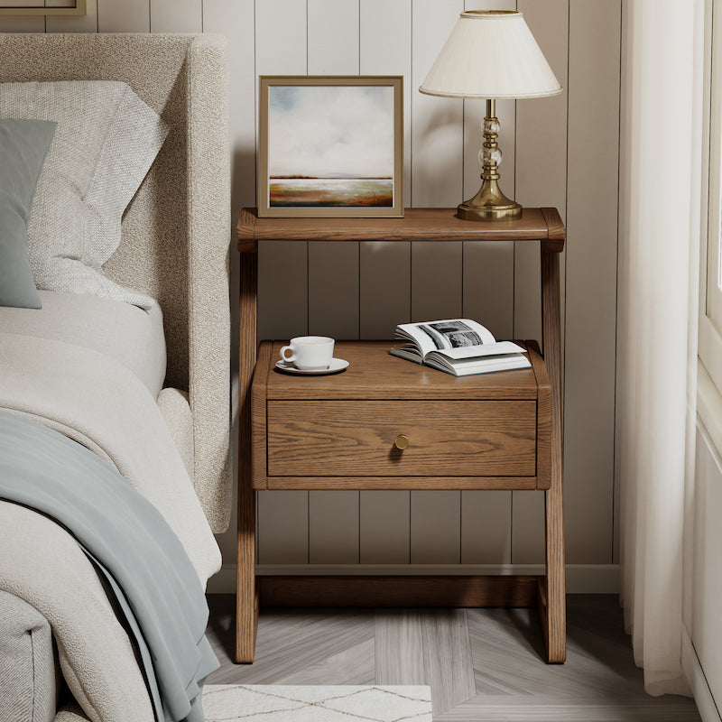 Wooden nightstand with a lamp, cup, and book next to a bed in a bedroom setting.